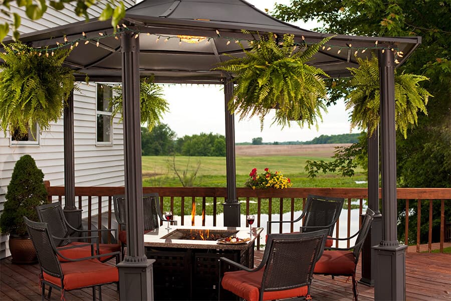A cozy gazebo on a deck surrounded by hanging ferns and seating, with a fire pit and scenic views of the landscape.