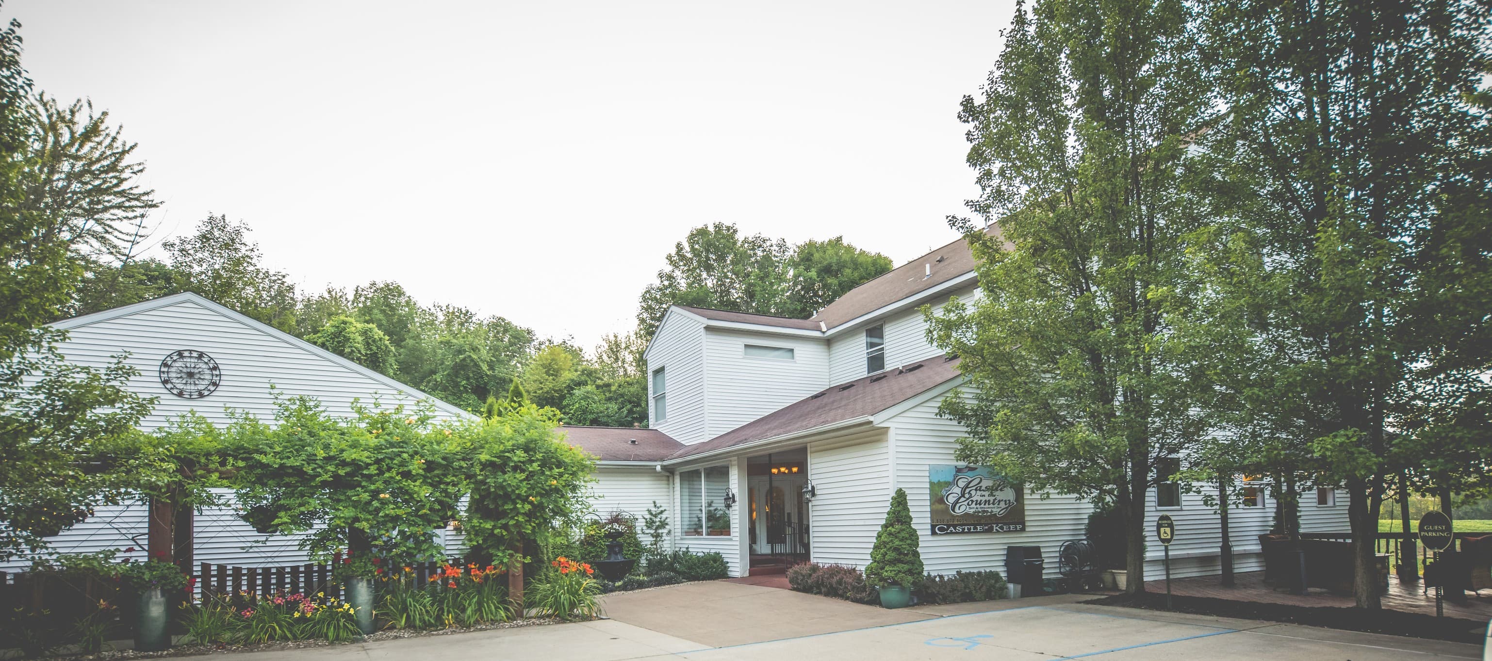 A white building with lush greenery and colorful flowers in front, set against a wooded backdrop.