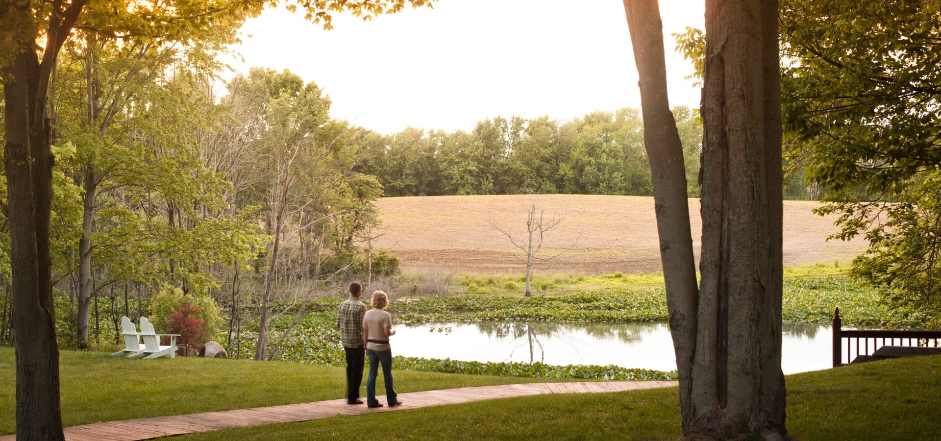 A couple stands on a path beside a tranquil pond, surrounded by lush greenery and fields.