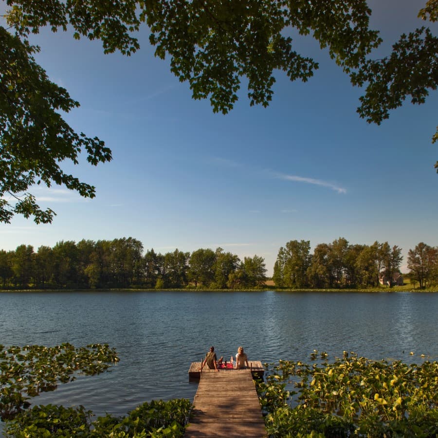 Two people sit on a wooden dock overlooking a tranquil lake surrounded by trees.
