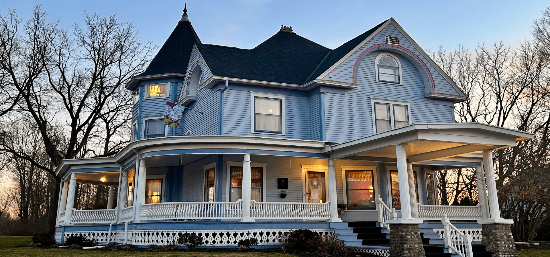 A large, ornate blue Victorian house with a wraparound porch and intricate roof details, surrounded by leafless trees at sunset.