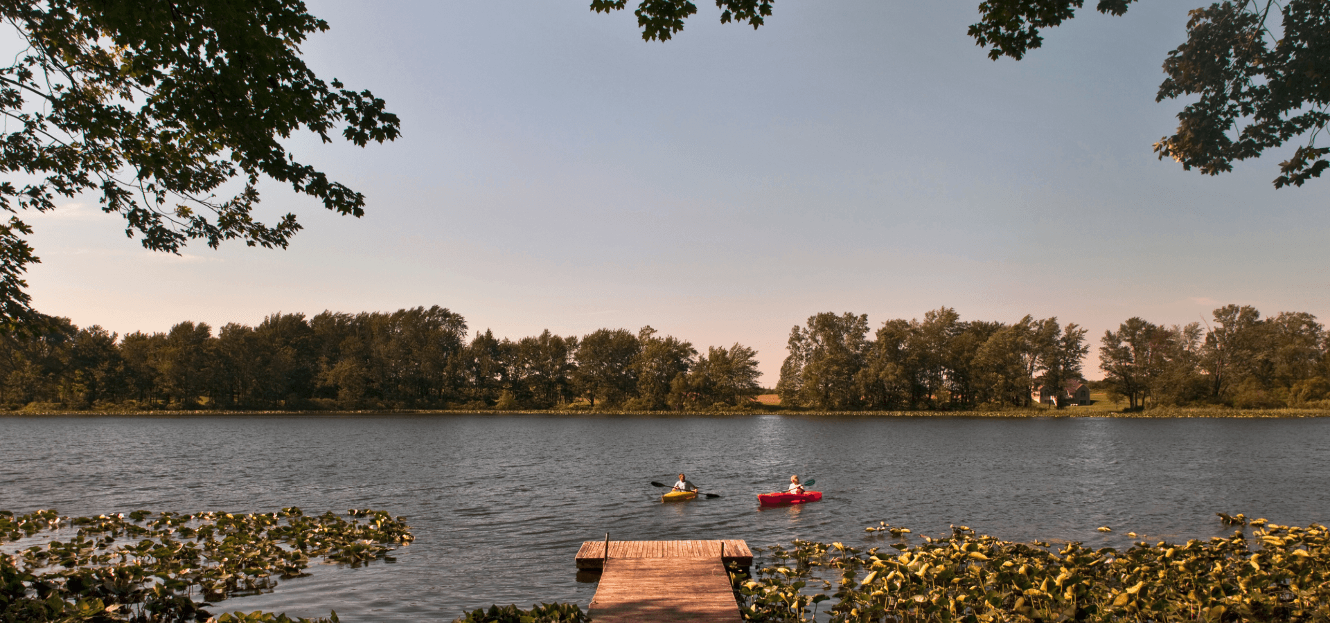 A tranquil lakeside scene with two kayakers near a wooden dock surrounded by lush greenery.