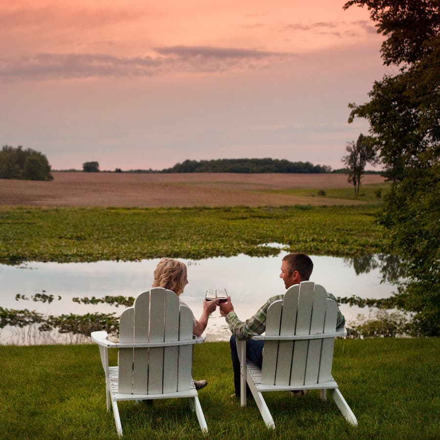 A couple toasts with glasses of wine while seated in lawn chairs by a tranquil pond at sunset.