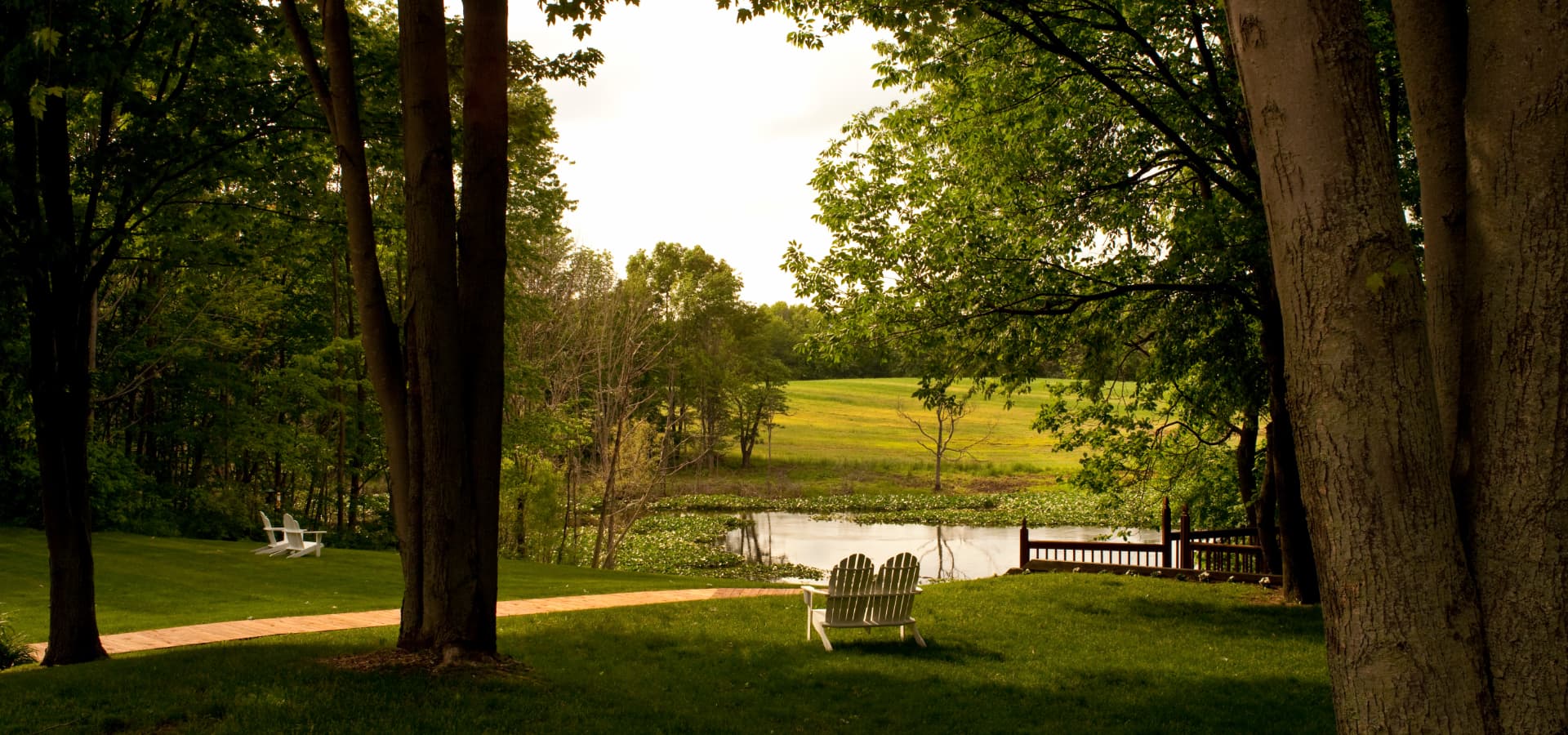 A serene view of a pond surrounded by lush greenery and an empty white lawn chair in the foreground.