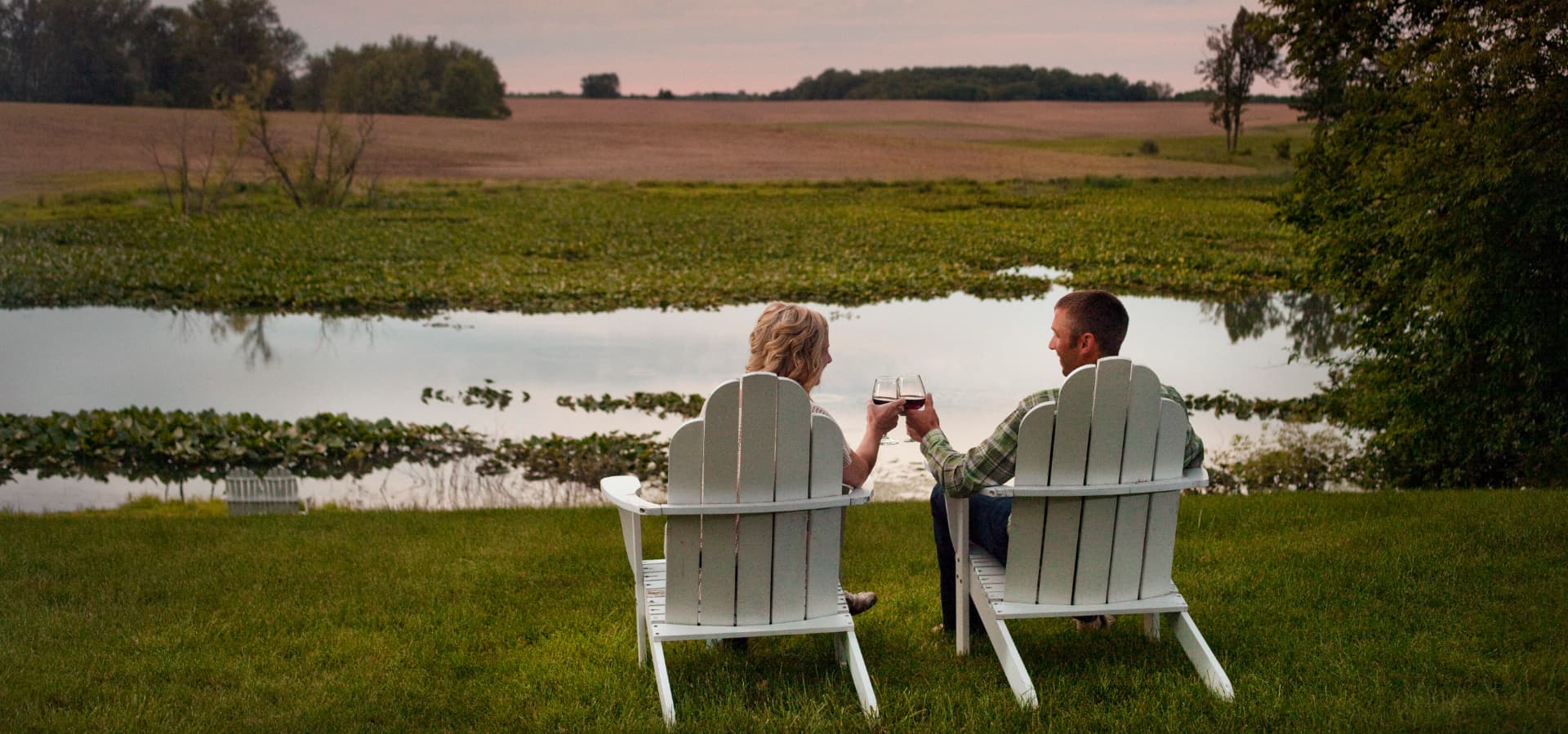A couple clinks glasses while sitting in white chairs by a serene pond at sunset.