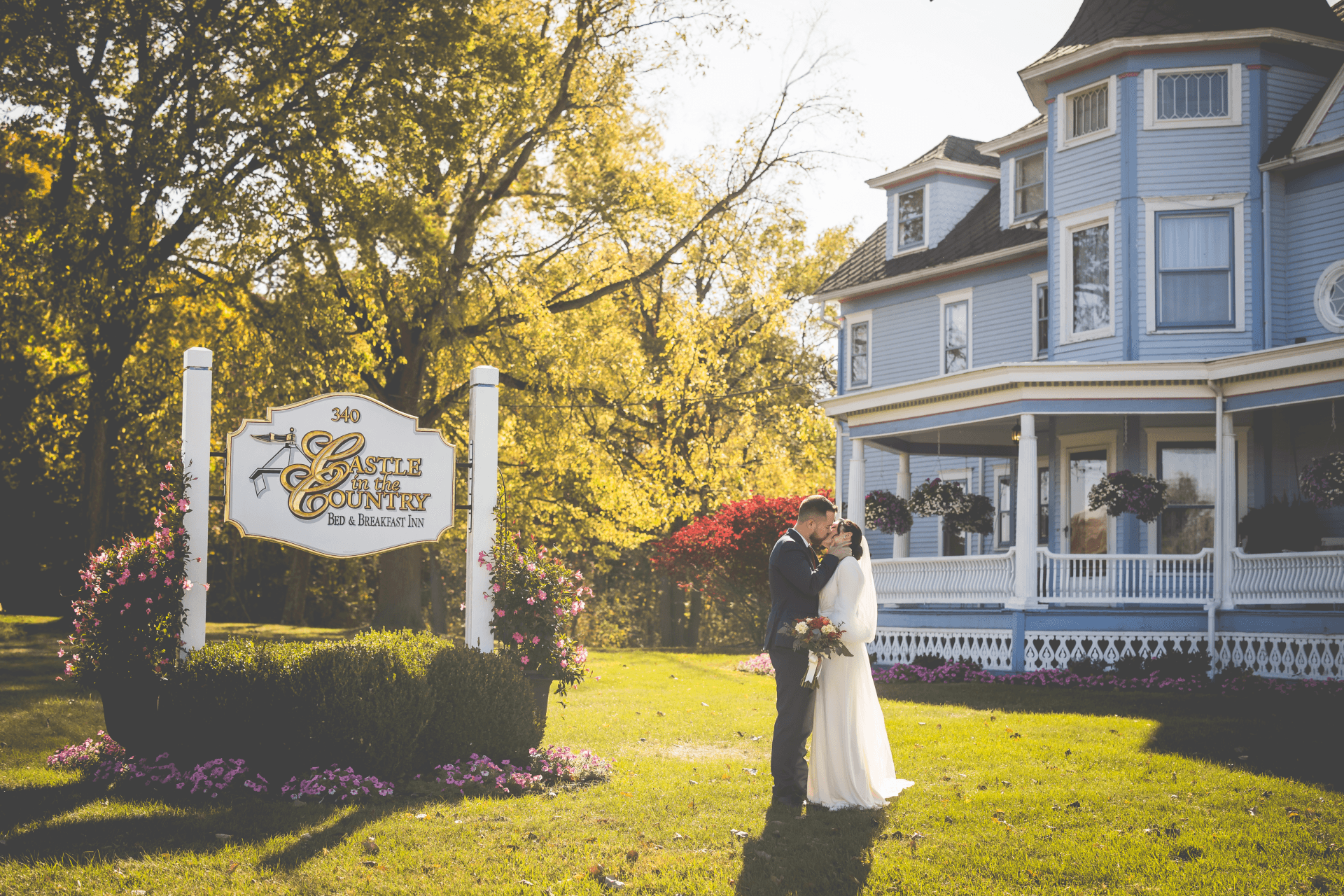 A couple embraces in front of a blue Victorian house with a welcoming sign and vibrant flowers.