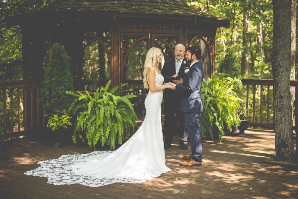 A bride and groom stand under a gazebo, exchanging vows with an officiant in a wooded setting.