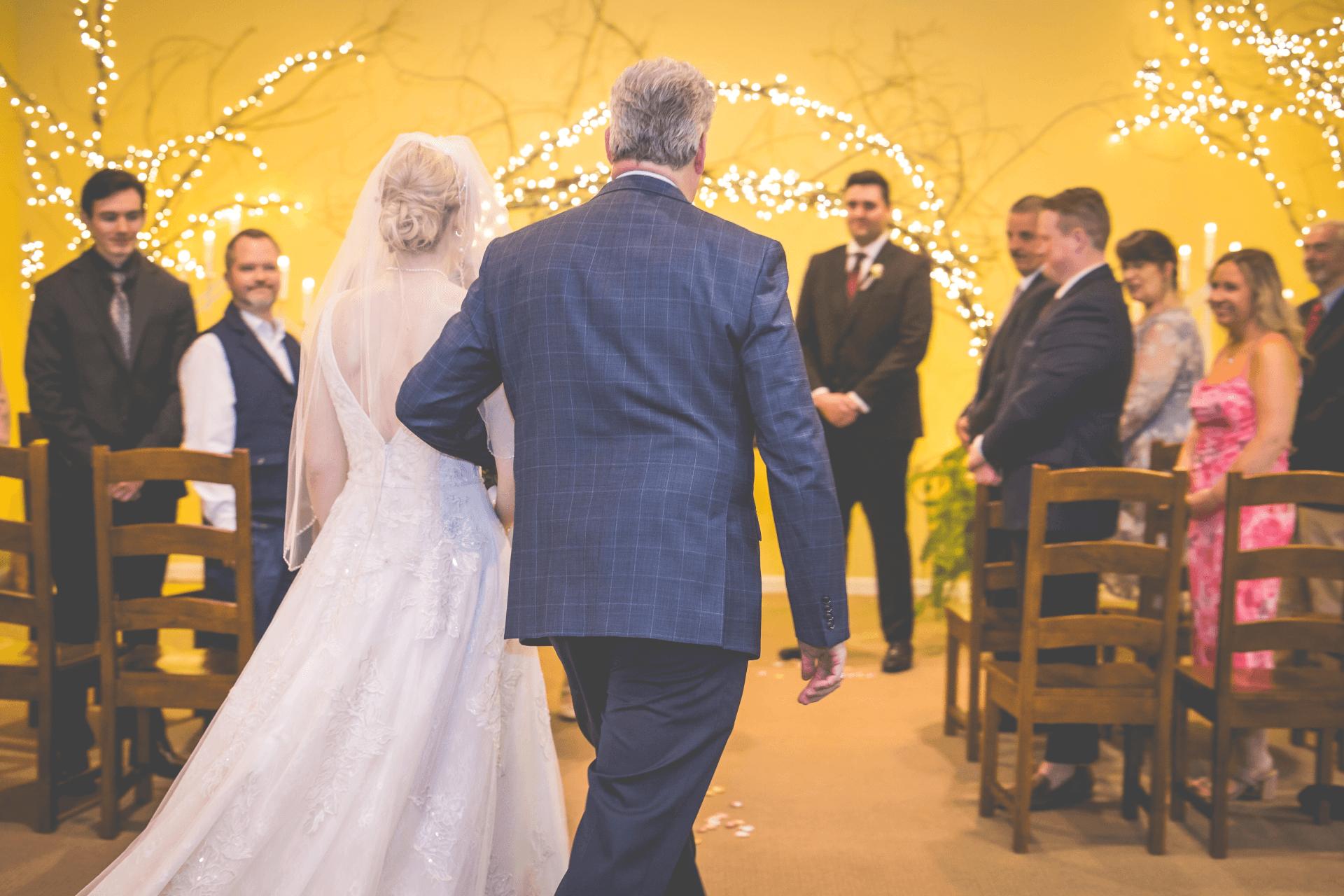 A bride walks down the aisle with her father, surrounded by guests in a warmly lit ceremony.