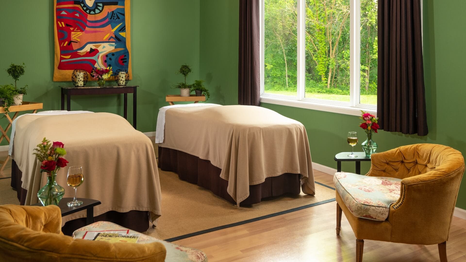 A soothing spa room with two massage tables, a cozy seating area, and large windows showcasing greenery outside.