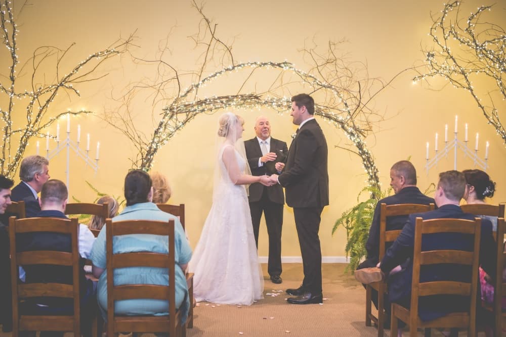 A bride and groom exchange vows during their wedding ceremony, with guests seated in front and a decorated arch in the background.