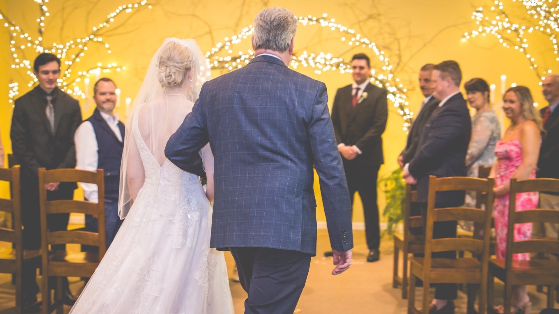 A bride, escorted by her father, walks down the aisle toward a wedding ceremony with guests seated around them.