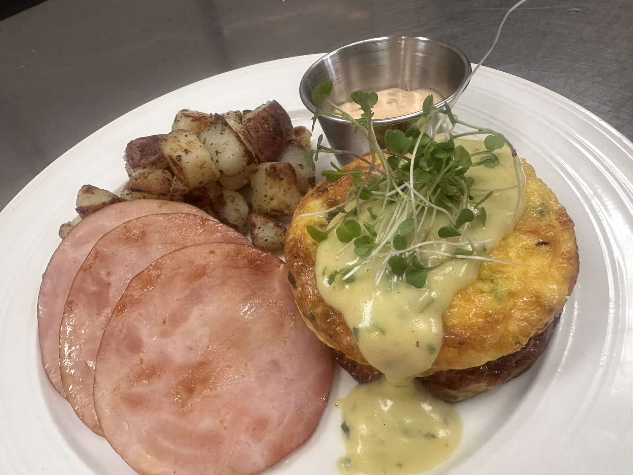 plate of royal egg and hollandaise sauce garnished with greens, slices of ham, and roasted potatoes
